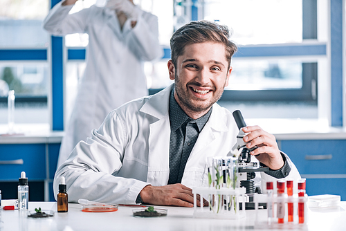 selective focus of happy biochemist near microscope and plants in test tubes