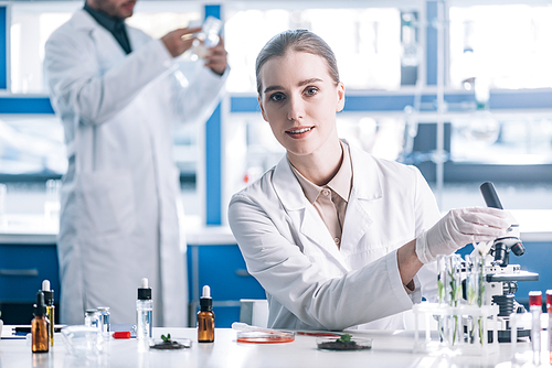 selective focus of happy and attractive biochemist near microscope and plants in test tubes