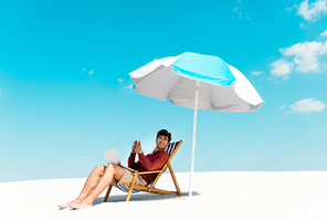 smiling freelancer sitting with laptop in deck chair under umbrella on sandy beach against blue sky