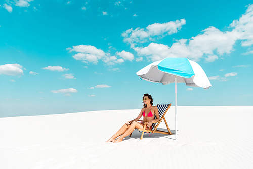 smiling beautiful sexy woman in swimsuit and sunglasses sitting in deck chair under umbrella on sandy beach