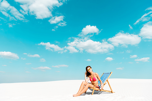 beautiful sexy 20대 여성 in swimsuit and sunglasses sitting in deck chair on sandy beach with blue sky and clouds