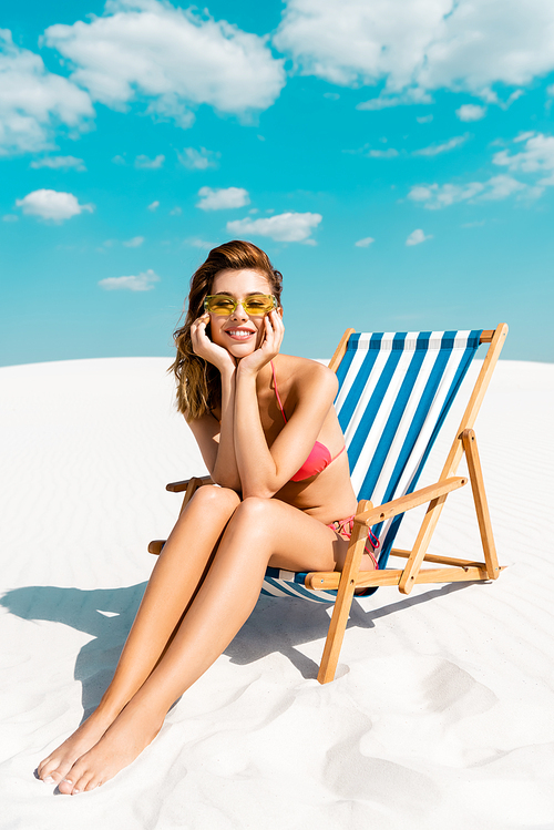 smiling beautiful sexy girl in swimsuit and sunglasses sitting in deck chair on sandy beach with blue sky and clouds