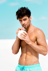 sexy man with muscular torso in swim shorts with coconut drink on sandy beach