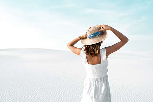 back view of beautiful girl in white dress and straw hat on sandy beach with blue sky