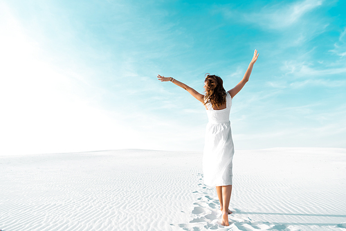 back view of beautiful girl in white dress with hands in air on sandy beach with blue sky