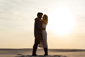 side view of young couple hugging on sandy beach at sunset