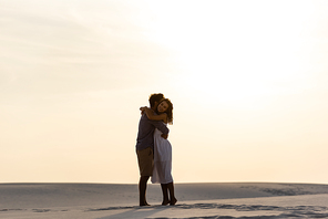side view of young couple hugging on sandy beach at sunset