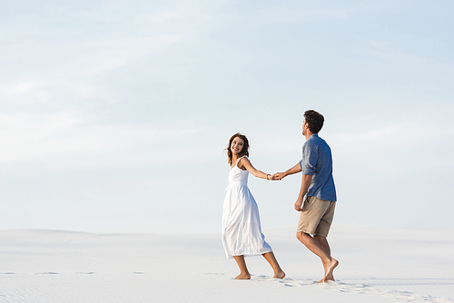 young couple walking on sandy beach and holding hands against blue sky