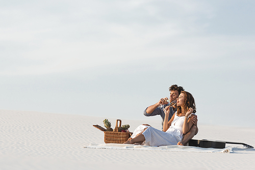 young couple drinking champagne while sitting on blanket with basket of fruits and acoustic guitar on beach