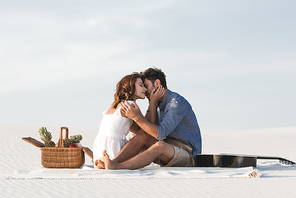 young couple hugging while sitting on blanket with basket of fruits and acoustic guitar on beach