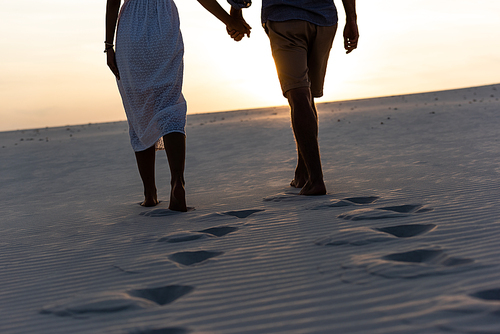 cropped view of man and woman holding hands while walking on beach against sun during sunset