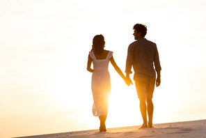 silhouettes of man and woman holding hands while walking on beach against sun during sunset