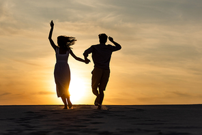 silhouettes of man and woman running on beach against sun during sunset