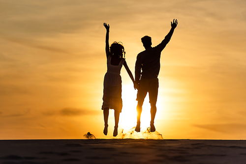 silhouettes of man and woman jumping on beach against sun during sunset