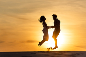 side view of silhouettes of man and woman jumping on beach against sun during sunset