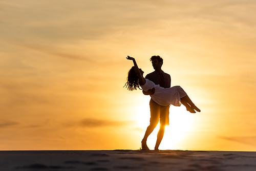 silhouettes of man spinning around woman on beach against sun during sunset