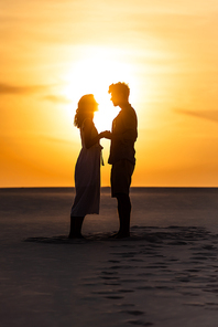 side view of silhouettes of man and woman holding hands on beach against sun during sunset