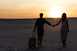 back view of couple walking on beach while holding hands with travel bag at sunset