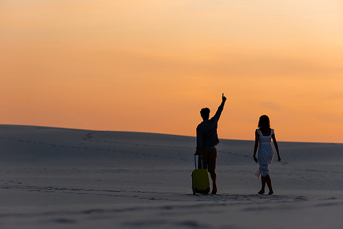 back view of couple walking on beach with raised hand and travel bag at sunset