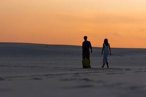 back view of couple walking on beach with travel bag at sunset