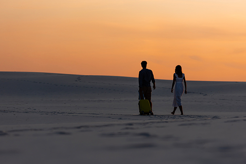 back view of couple walking on beach with travel bag at sunset
