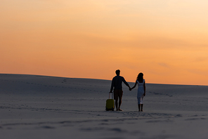 back view of couple walking on beach while holding hands with travel bag at sunset