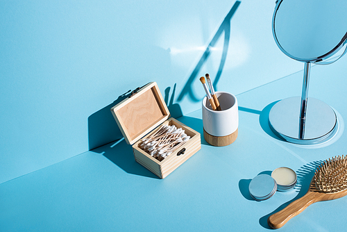 Toothbrush holder with cosmetic brushes, box of ear sticks, mirror, jar of wax and hair brush on blue background, zero waste concept