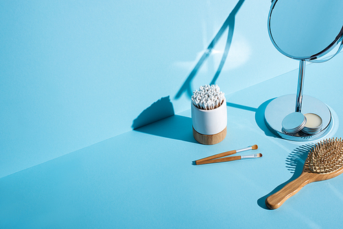 Ear sticks in toothbrush holder near cosmetic brushes, mirror with jar of wax and  hair brush on blue background, zero waste concept