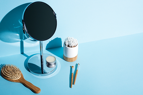 Mirror with jar of wax next to hair brush, cosmetic brushes, toothbrush holder with ear sticks on blue background, zero waste concept