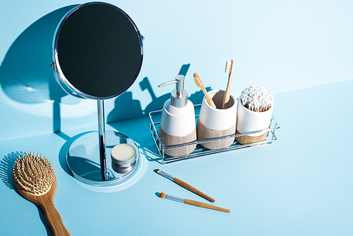 Toothbrush holders with hygiene items, dispenser on bathroom shelf with mirror, hair brush, cosmetic brushes on blue background, zero waste concept