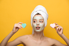 surprised girl with nourishing facial mask holding heart-shaped cosmetic sponges and looking away on yellow background