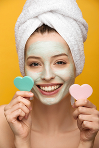 cheerful girl with nourishing mask on face holding heart-shaped cosmetic sponges isolated on yellow