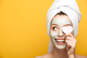 cheerful girl with nourishing facial mask covering eye with heart-shaped cosmetic sponge on yellow background