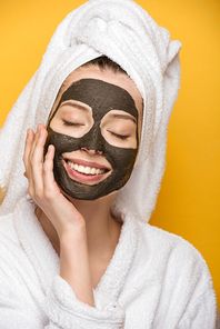 happy girl with facial clay mask and towel on head touching face isolated on yellow