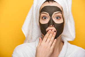 shocked girl with facial clay mask and towel on head covering mouth with hand isolated on yellow