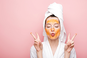 young woman with citrus facial mask showing victory gestures isolated on pink