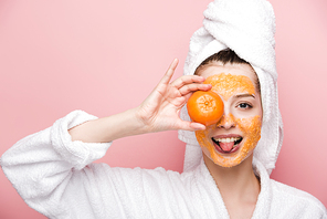 cheerful girl with citrus facial mask covering eye with tangerine and sticking out tongue isolated on pink