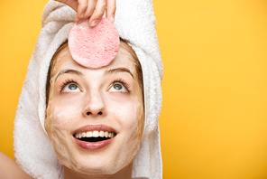 cheerful girl with moisturizing facial mask looking at cosmetic sponge isolated on yellow