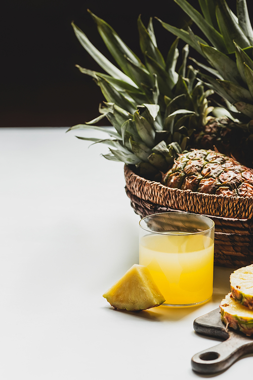 fresh pineapple juice in glass near delicious fruit in basket on white surface isolated on black