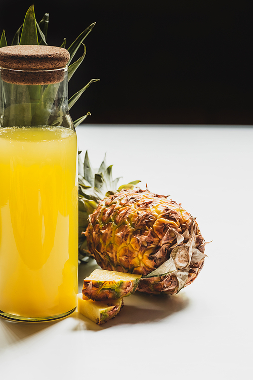 fresh pineapple juice in bottle near cut delicious fruit on white surface isolated on black