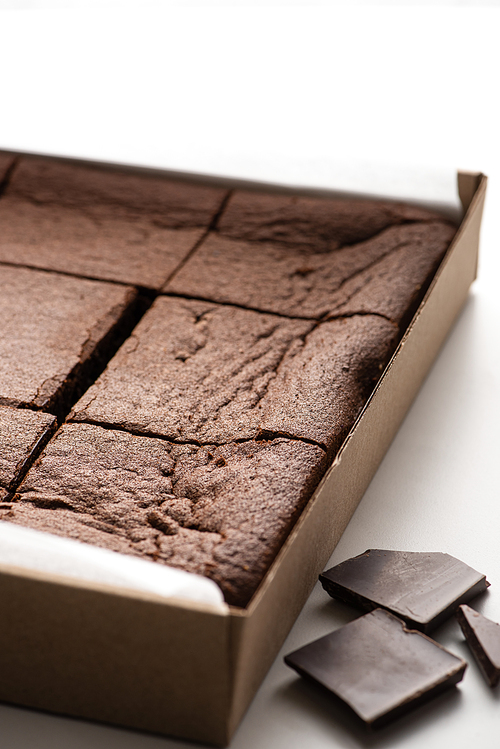 close up view of delicious brownie pieces in cardboard box near dark chocolate on white background