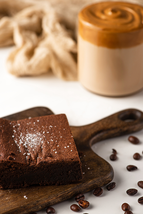 selective focus of delicious brownie piece on wooden cutting board with coffee beans on white background