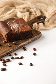 delicious brownie piece on wooden cutting board with coffee beans near cloth on white background