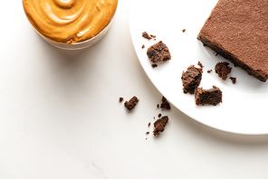 top view of delicious brownie piece on plate near peanut butter on white background