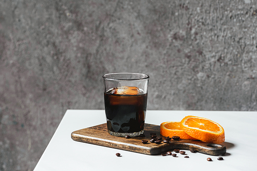 cold brew coffee with ice in glass near orange slices on chopping board and coffee beans on white table