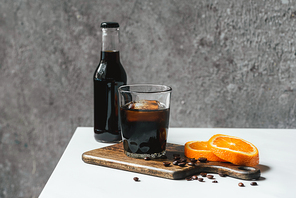 cold brew coffee with ice in glass and bottle near orange slices on chopping board and coffee beans on white table