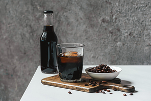 cold brew coffee with ice in glass and bottle near coffee beans on chopping board on white table