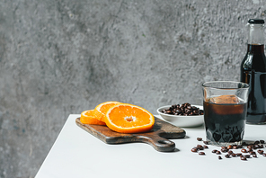 cold brew coffee with ice in glass and bottle near orange slices on chopping board and coffee beans on white table
