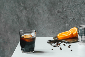 selective focus of cold brew coffee with ice in glass near orange slices on chopping board and coffee beans on white table