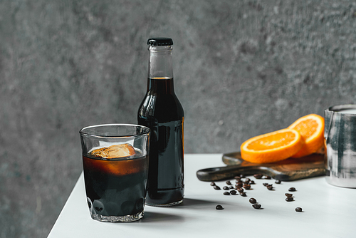 selective focus of cold brew coffee with ice in glass and bottle near orange slices on chopping board and coffee beans on white table